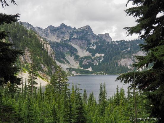 A rocky ridgeline with multiple peaks under a cloudy sky, above a quiet lake, all framed by conifer branches