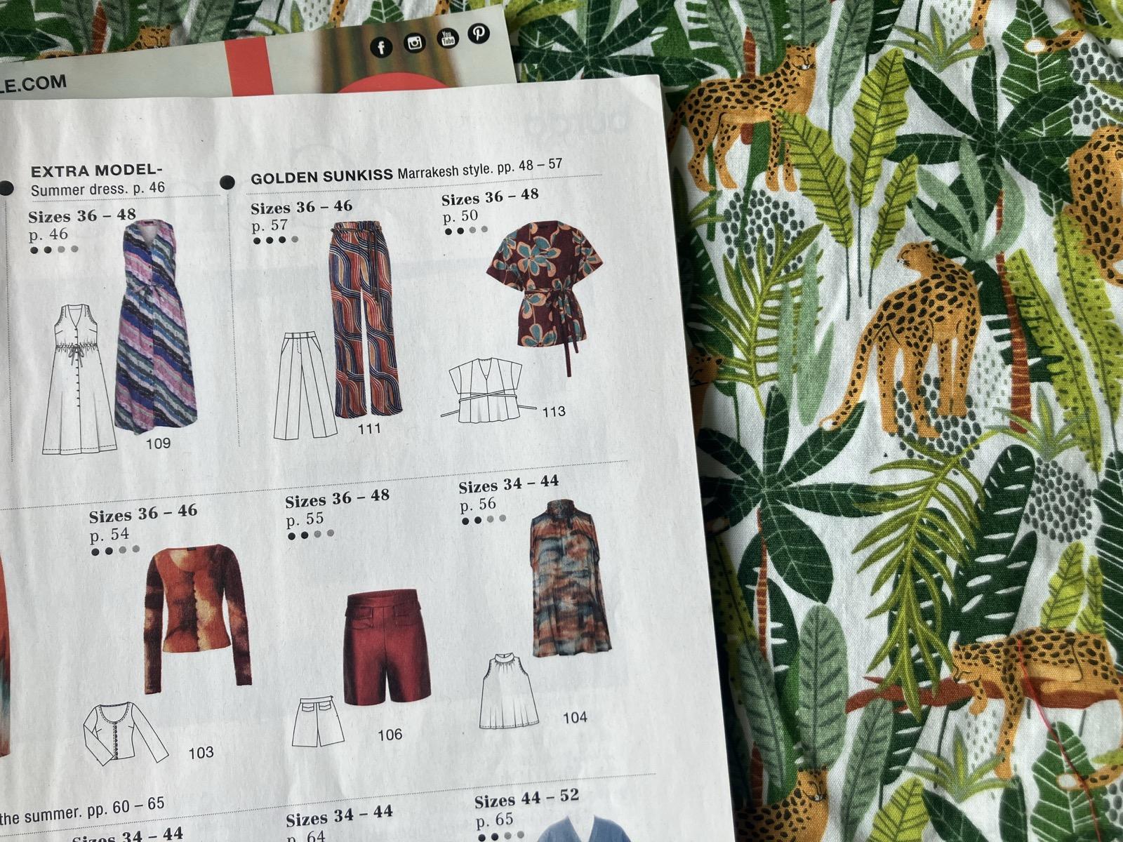 Photo of fabric with a green jungle plant print with leopards amongst the plants. Sitting on top is a magazine page showing a long sleeveless shirt dress, a pair of trousers, 3 tops, and a pair of shorts