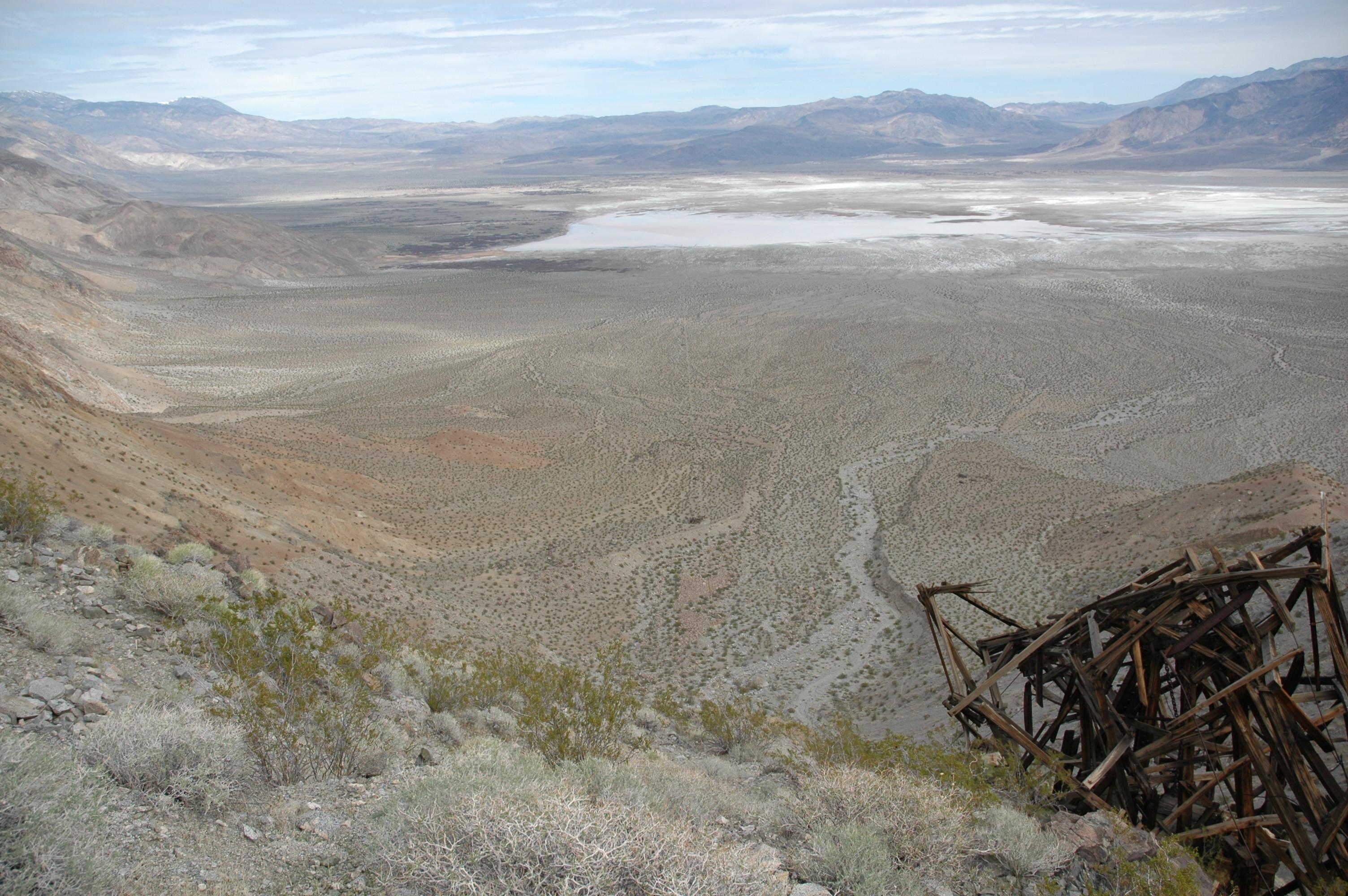 A deep desert valley replete with dry salt beds and visible sandy washes is seen below from a vantage point on a high ridge in the mountains above. An old collapsing wooden structure is visible in the foreground on the right.