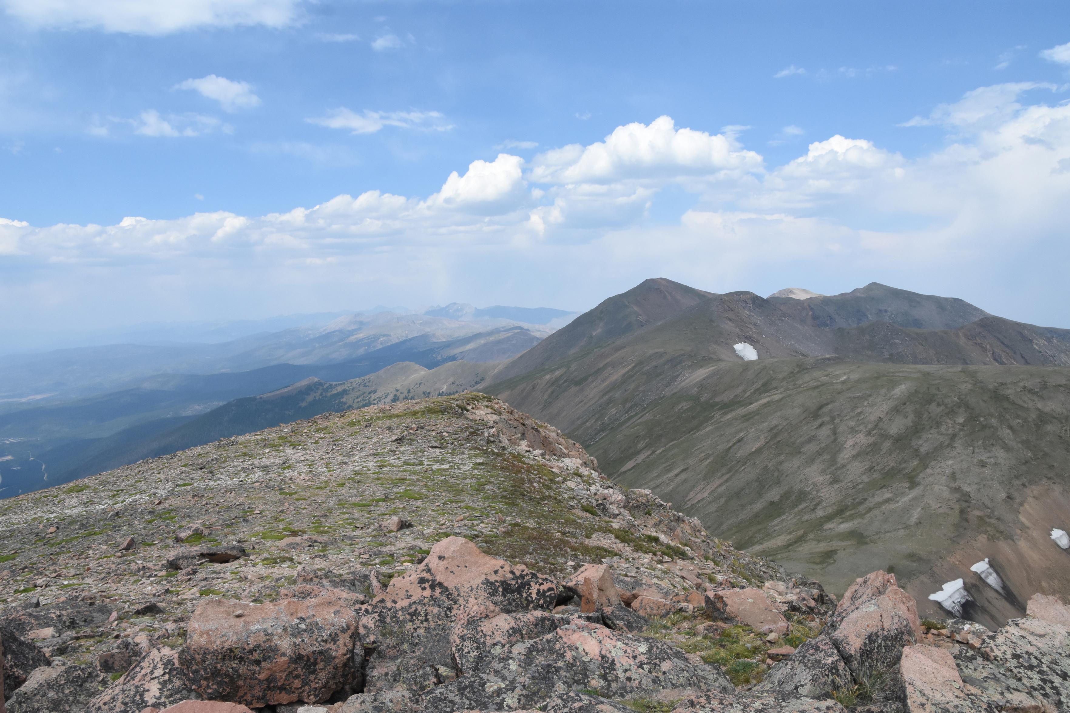 A series of high peaks continues North toward the distant horizon. Rocky alpine tundra in the foreground drops off to a wide valley to the West.