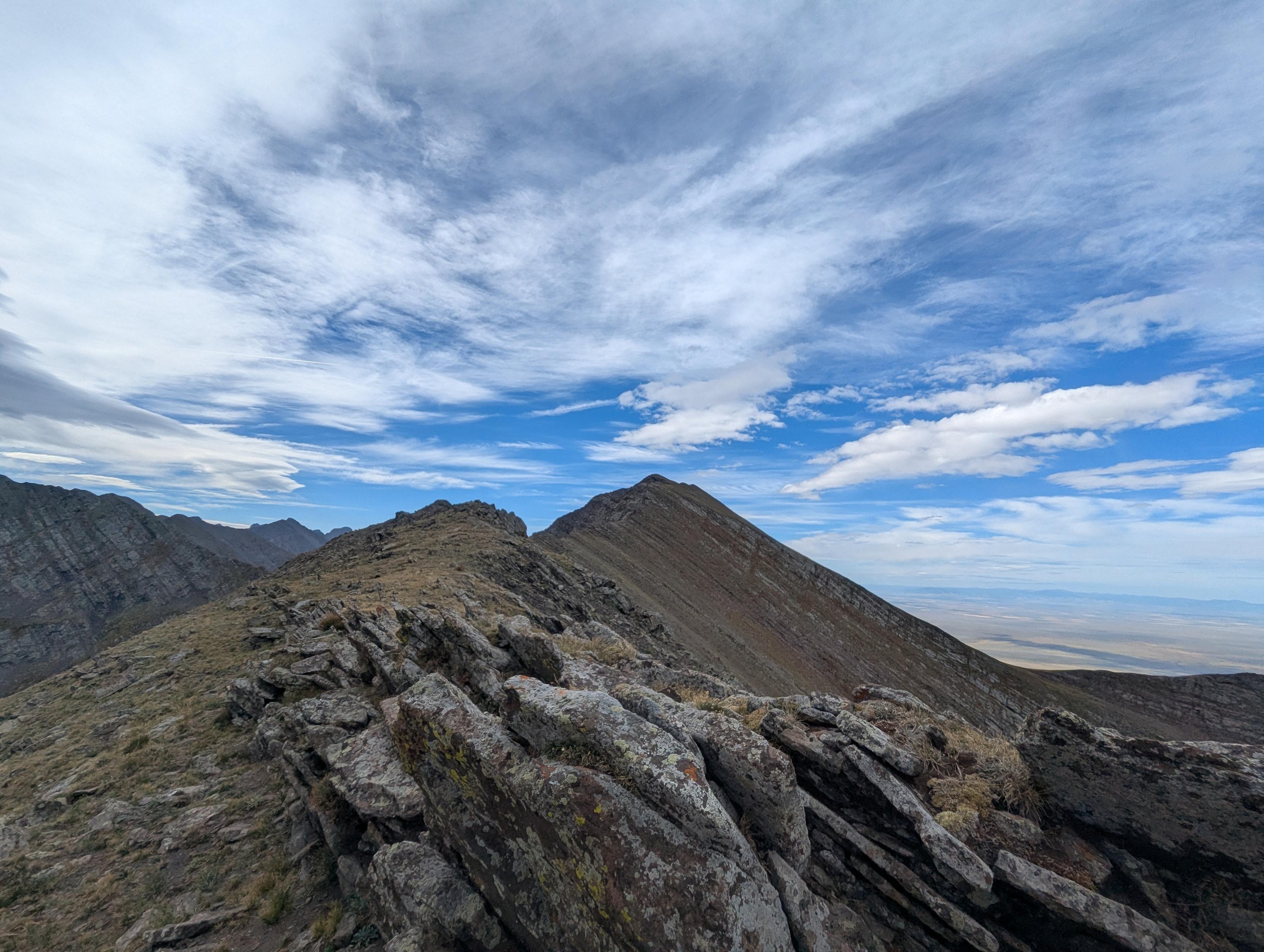 A ridge of rocks like a knife edge curves toward a pointy mountain peak. Blue sky and wispy clouds drift over the mountain landscape. A distant valley is visible on the horizon thousands of feet below.