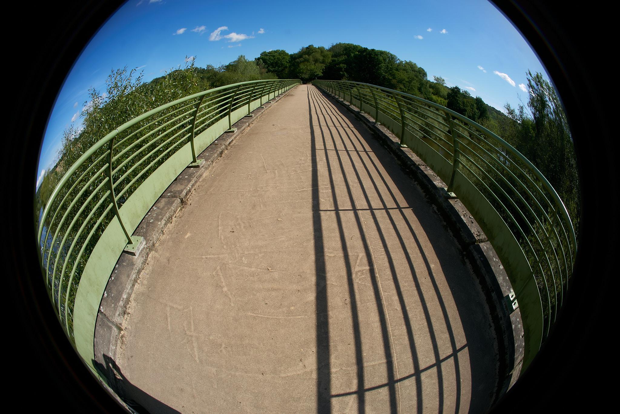 Wide Angle shot of a footbridge over a river with no river in view.  The angle is a wide fish eye shot with the bridge taking up most of the image, the shadow of the railings run directly, the shadow of the photographer is in view on the bottom left.
